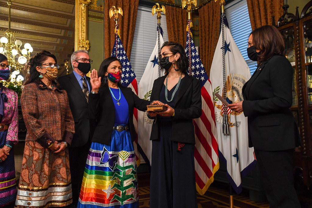 Secretary Deb Haaland in a bright blue dress with a rainbow-striped skirt swearing into her position next to Vice President Kamala Harris.